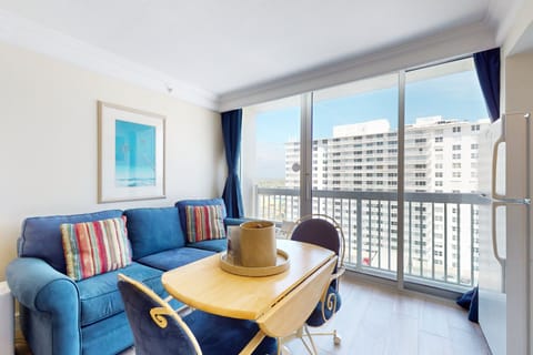 Relax in this beachy living space featuring a cozy blue sofa, vibrant décor, and floor-to-ceiling windows leading to a private balcony with ocean views—perfect for your seaside getaway.