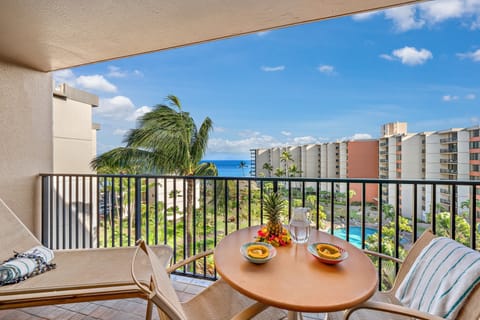 Private balcony with a round wooden table set for two, offering panoramic views of resort buildings, lush greenery, and the nearby ocean under a bright blue sky.