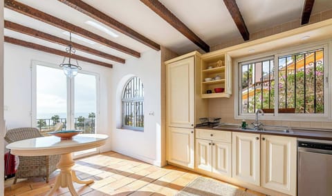 Bright Mediterranean kitchen with terracotta floors, wooden beams, cream cabinets, and a round table by glass doors opening to ocean views, filled with natural light and coastal charm.
