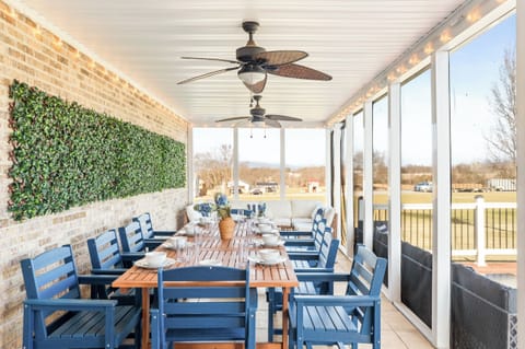 Screened‑in porch with a long farmhouse table, string lights, and ceiling fans—perfect for breezy outdoor dinners, game nights, and sunrise coffee with peaceful country views.