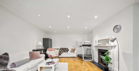 This bright, modern living room features white walls, herringbone floors, and a classic fireplace. It blends relaxation and fitness with white sofas, a matching coffee table, and a corner treadmill.