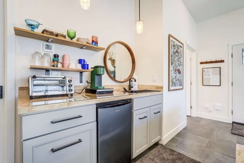 The kitchenette features open wood shelving with dishes and glassware, a toaster oven, Keurig machine, compact fridge, and a round wall mirror, all set against clean cabinetry and warm overhead lighting.