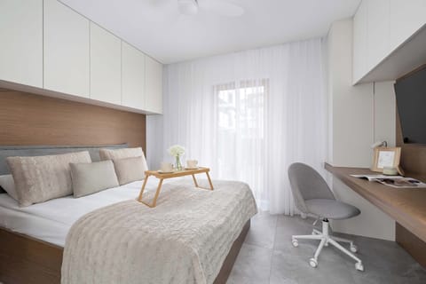 A bright bedroom with a comfortable double bed, soft textiles, and a neutral color palette. A compact desk with a chair creates a practical workspace by the window.