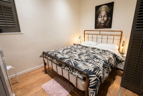 The double bedroom features a copper-framed bed dressed in a monochrome leaf-print duvet, flanked by bedside lamps and plantation shutters on two sides.