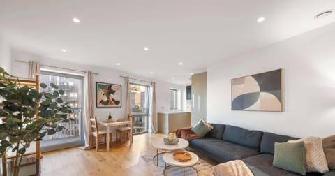 A bright, modern living room with light wood floors and white walls, featuring a large navy sectional and contemporary art. Floor-to-ceiling windows and recessed lighting create an airy, well-lit space.