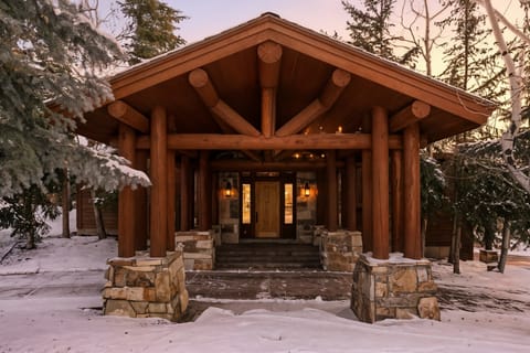 Exterior view of a luxury log cabin with snow-covered trees and warm interior lighting, showcasing rustic mountain architecture.