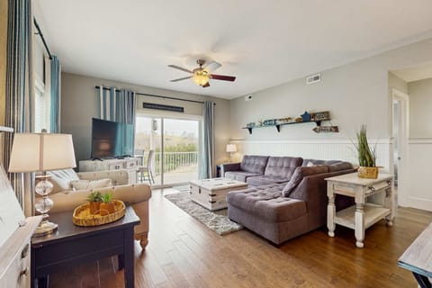 Bright living room with sectional sofa, armchair, TV, coffee table, and sliding glass doors leading to the balcony.