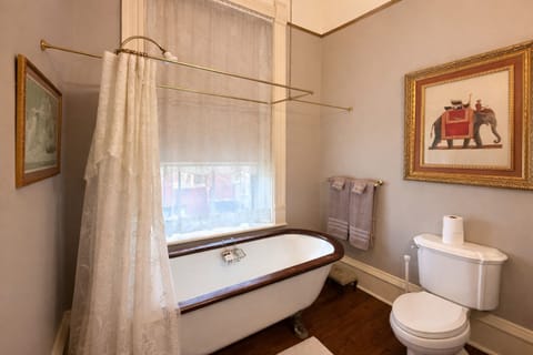 Clawfoot bathtub with wood-trimmed rim, lace curtain, towel bar, and historic wall art.