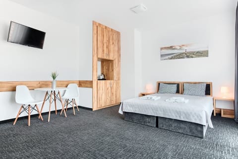 A bright living area featuring a two beds, a small table with two chairs, and wooden accents along the wall. A mounted TV and artwork complete the minimalist decor.

