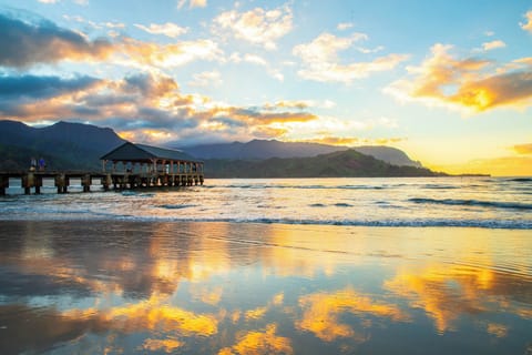 Golden sunset paints the sky over pristine beach waters, with a historic pier and dramatic mountain backdrop creating an unforgettable Hawaiian paradise scene.