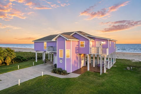 Aerial sunset view of a colorful Galveston beachfront vacation home with direct Gulf views, spacious lawn, elevated construction and generous paved parking.