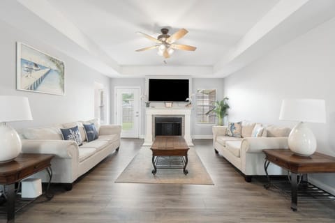 Step into comfort in this spacious living room, featuring matching sofas, a stylish ceiling fan, and a cozy fireplace as the centerpiece. Natural light pours in from multiple windows and a glass door, creating the perfect setting for relaxation.