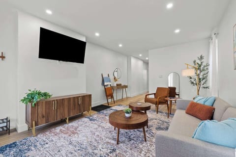 Modern living room featuring a comfortable sofa, warm wood-toned furniture, and a large 55-inch Roku Smart TV for all your streaming needs. The space is brightened by recessed lighting, a colorful area rug, and stylish mid-century accents.
