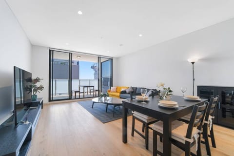 Open plan living and dining with a grey corner sofa and a yellow armchair, a TV on a dark console, and a round stone top coffee table. Sliding glass doors open to the balcony and a potted plant sits on the TV console.