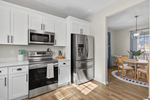 A bright, modern kitchen features white cabinetry, stainless-steel appliances, and warm wood flooring. The open layout flows directly into the dining and living spaces. 