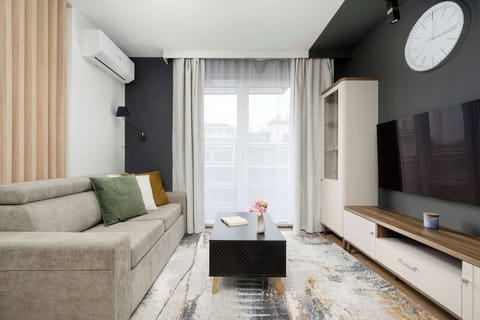 A modern living room with a gray accent wall, wall-mounted TV, and light-colored sofa. A small coffee table sits on a rug, creating a cozy seating area. Natural light enters through large windows with curtains.