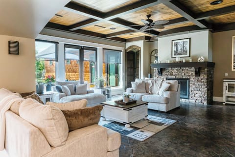Step into a cozy living room with a stone fireplace crackling at the heart of it. Gray armchairs by the fire, large windows with mountain views, and rustic décor create a warm, inviting space.