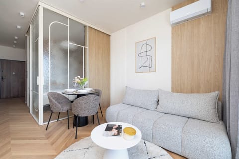 A bright and modern studio living area with a light sofa, soft decor, and a partially visible glass-walled shower.