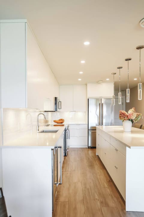 A modern kitchen with white cabinets, light countertops, stainless steel fridge, and a spacious island with bar stools and pendant lights.
