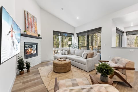 Unwind in your bright living space with cozy fireplace, plush seating, and forest views streaming through panoramic windows.