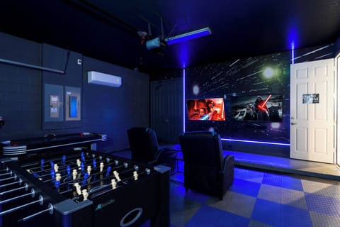 Welcome to our lively game room, where fun awaits with a foosball table and a large screen for movies. The vibrant blue lighting against the sleek black walls sets the perfect mood for relaxation and entertainment.