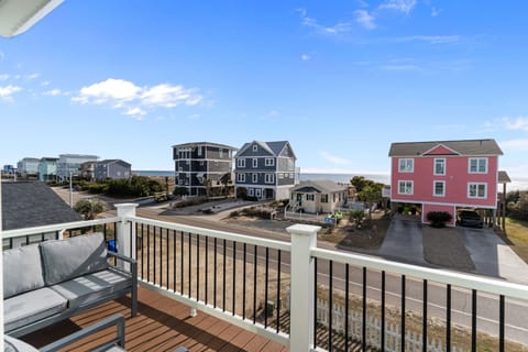 Balcony with cushioned seating and black railing overlooking the road, colorful beach houses, dunes, and Atlantic Ocean beneath a bright blue sky.