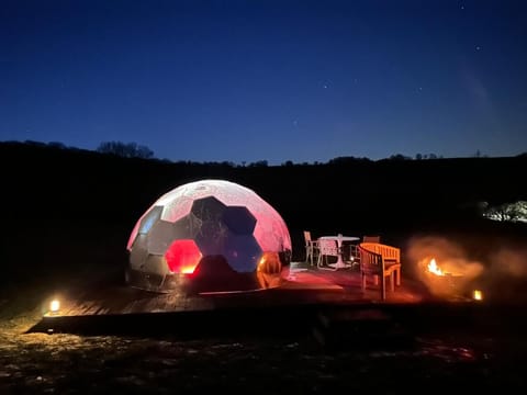 A lit geodesic dome sits on a raised deck with outdoor seating and a fire pit under the night sky