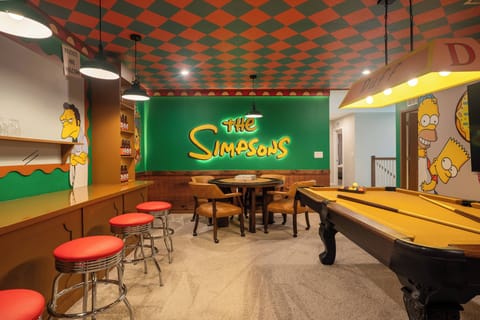Step into a vibrant game room featuring playful "The Simpsons" murals and a striking checkerboard ceiling. With cozy red barstools and a classic pool table, this space invites lively gatherings and friendly competition.