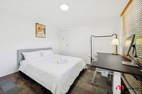 Comfortable bedroom featuring a queen-size bed, bedside table with lamp, study desk, and large window with blinds.