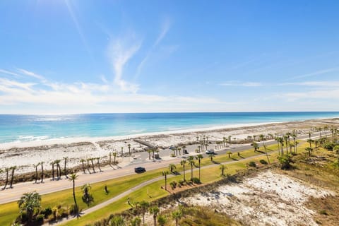 Stunning aerial view of pristine white sand beach with turquoise waters, palm-lined coastal road, and manicured green spaces.