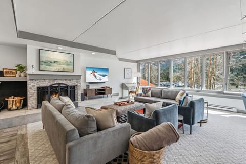 Sink into plush seating by your crackling fireplace, where floor-to-ceiling windows frame stunning winter forest views in this bright, modern living room.