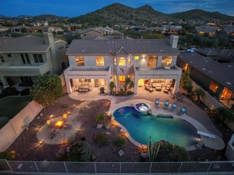 Aerial view of a luxurious desert retreat featuring a sparkling pool, hot tub, and expansive outdoor entertaining spaces set against scenic mountain foothills at twilight.