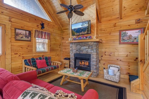 Cozy up in your rustic cabin living room featuring a crackling stone fireplace, warm wood walls, and mountain views on TV.