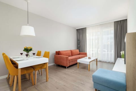 A bright living and dining area with a wooden table, mustard-yellow chairs, a salmon-colored sofa, and large curtains letting in daylight.