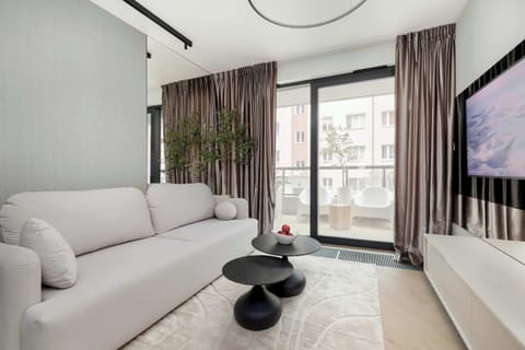 A bright living room with a large light-colored sofa facing a wall-mounted TV. Floor-to-ceiling windows bring in natural light and connect the space to a balcony. Neutral tones and soft textures create a calm atmosphere.