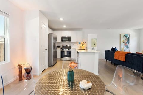 Bright kitchen and dining area featuring stainless-steel appliances, wide countertops, and a modern round table overlooking the backyard. The open layout flows into the living room, where a 55” TV creates the perfect place to relax after meals.