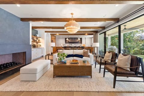This sophisticated living room showcases exposed wood beams, a striking fireplace, and expansive glass doors framing lake views. Plush seating, curated décor, and a statement chandelier create an elegant yet inviting atmosphere.