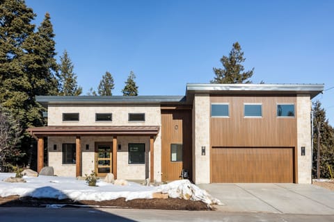 Striking exterior of this modern mountain retreat, showcasing clean architectural lines, stone and wood accents, a private garage, and a welcoming covered entry—all set against a peaceful alpine backdrop.
