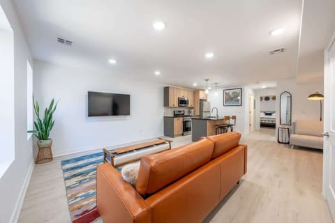 An open living area with a leather sofa, textured rug, and wall-mounted TV. The space flows naturally into a modern kitchen, creating a bright and connected environment for relaxing.