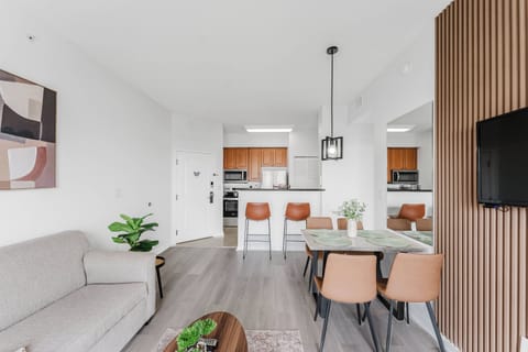 Experience a stylish living area with a cozy gray sofa and warm wooden accents, creating a welcoming atmosphere. The modern dining table and chairs invite you to enjoy meals, all illuminated by natural light from the kitchen.