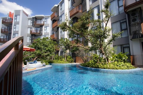 Large outdoor swimming pool surrounded by tropical plants and modern apartment buildings.