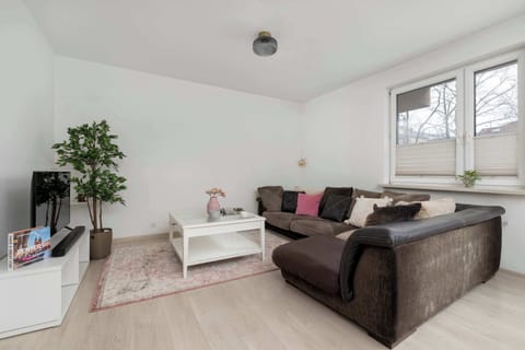 A bright living room with a brown sectional sofa and light wood flooring. White walls and soft décor create a calm, welcoming atmosphere. A small plant adds a natural accent.