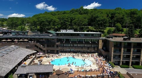 The outdoor pool area bustles with life under a stunning azure sky, as guests bask in the warm sun with the majestic forest backdrop, perfect for summer fun.