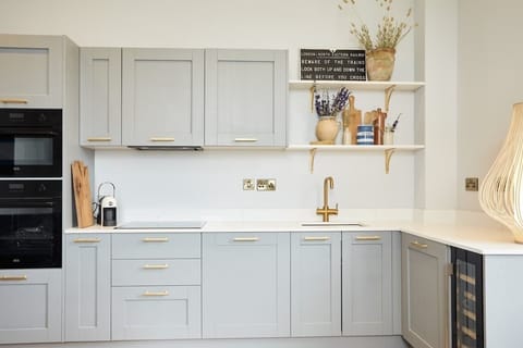 A sleek kitchen with gray cabinets, white counters, and built‑in oven. Gold faucet, floating shelves, and decorative touches create a warm, welcoming space filled with natural charm.