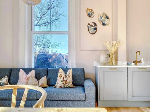 Natural light fills a cozy lounge with blue sofa and textured cushions. A modern kitchenette with gold fixtures and stylish cabinetry adds convenience, while abstract wall art enhances the inviting atmosphere.