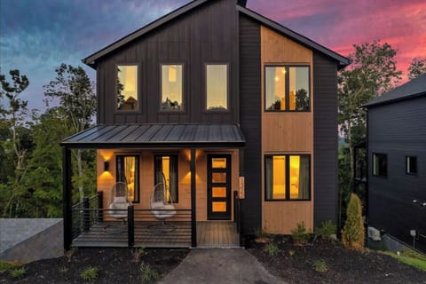 Modern two-story home with striking black and wood exterior featuring warm interior lighting against a dramatic sunset sky.