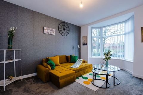 Living room view showing the mustard sofa, glass coffee table, and bay window with vertical blinds.