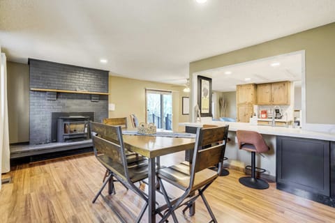 Welcome to Ravine Abode! The dining area features a high-top wooden table with seating for four, set against a dark grey brick fireplace. Light hardwood flooring and neutral walls create a bright, open atmosphere that connects to the nearby kitchen.