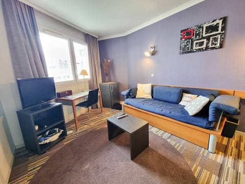 Salon avec canapé lit, coin TV, Box wifi, coin bureau, table basse et réfrigérateur.