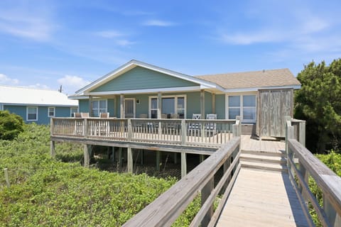 Charming coastal beach house with an elevated deck and private boardwalk access, nestled among natural dunes and greenery under sunny skies.